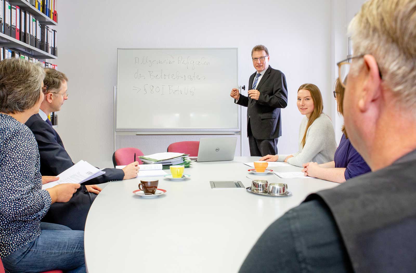 Ein Seminarraum: Rechtsanwalt Klaus Pahde erklärt etwas an einem Whitebord. Um einen großen ovalen Tisch herum sitzen zwei Männer und drei Frauen, die ihm zuhören.
