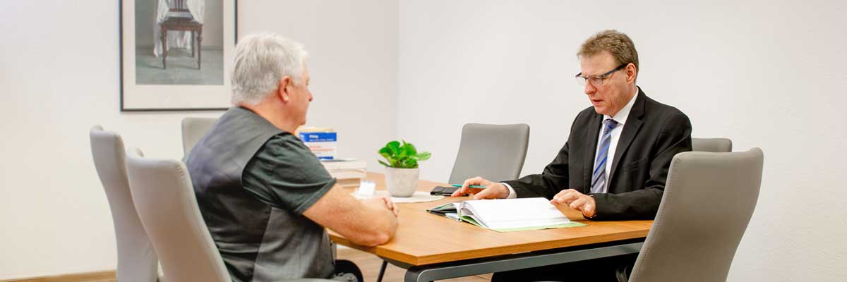 Zwei Herren sitzen sich an eiem Arbeitstisch in einem neutral gehaltenen Büro gegenüber.